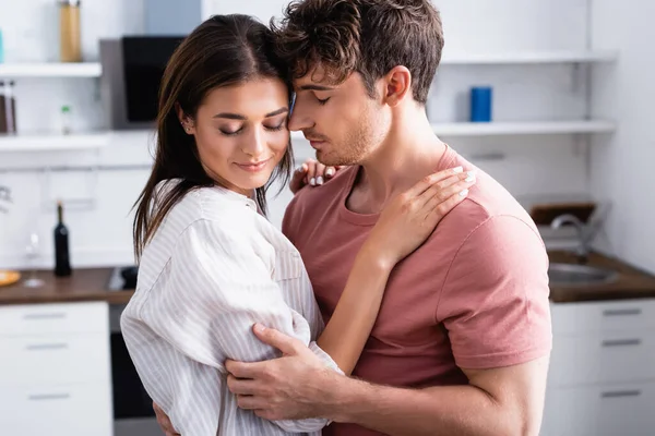 Joven abrazando novia con los ojos cerrados en la cocina - foto de stock