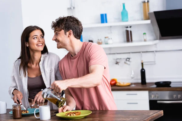 Sorrindo homem derramando chá perto da namorada cortando waffle na mesa da cozinha — Fotografia de Stock