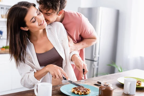 Homem beijando sorridente namorada cortando waffle perto de propagação de chocolate e copos na mesa — Fotografia de Stock