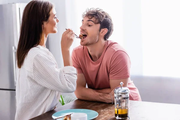 Smiling woman feeding boyfriend with waffle near tea on blurred foreground — Stock Photo