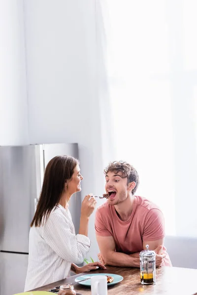 Femme gaie nourrissant petit ami avec gaufre près du thé pendant le petit déjeuner dans la cuisine — Photo de stock