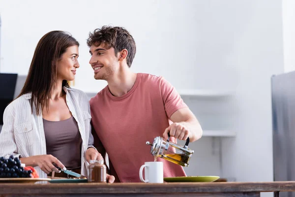 Hombre sonriente mirando a su novia mientras vierte té cerca de frasco de chocolate extendido en primer plano borroso - foto de stock