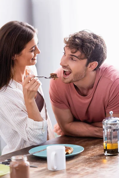 Smiling woman holding fork with waffle near boyfriend, tea and chocolate spread on blurred foreground — Stock Photo
