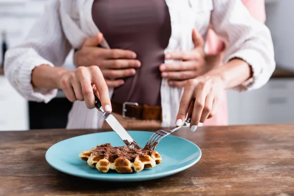 Cropped view of woman cutting waffle with chocolate spread near boyfriend hugging on blurred background — Stock Photo