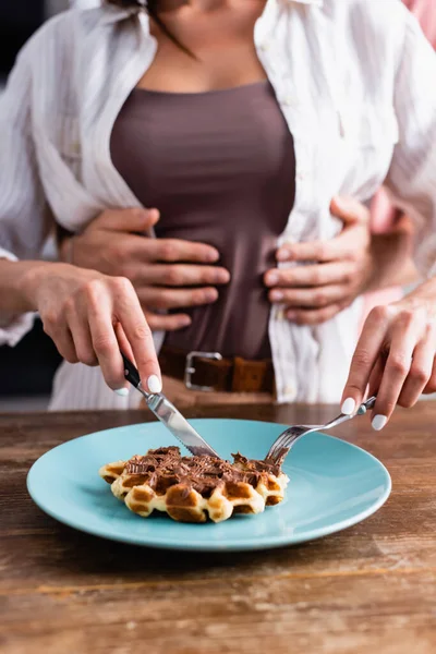 Vista cortada de jovem mulher cortando waffle saboroso perto do namorado abraçando no fundo borrado — Fotografia de Stock