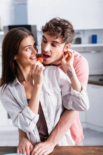 Mulher alegre alimentando namorado com morango fresco na cozinha — Fotografia de Stock