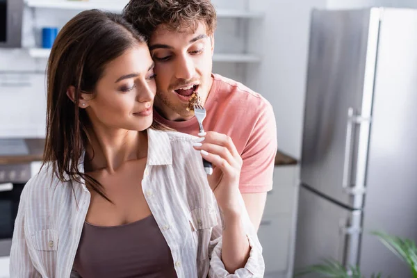 Young woman feeding boyfriend with piece of waffle in kitchen — Stock Photo