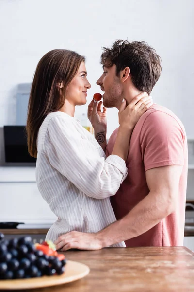 Side view of smiling woman hugging and feeding boyfriend with strawberry near grape on blurred foreground — Stock Photo