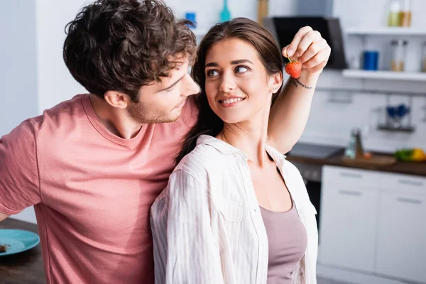 Young man holding strawberry near smiling girlfriend in kitchen — Stock Photo