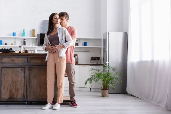 Hombre abrazando novia con los ojos cerrados cerca de uva y té en la mesa de la cocina - foto de stock