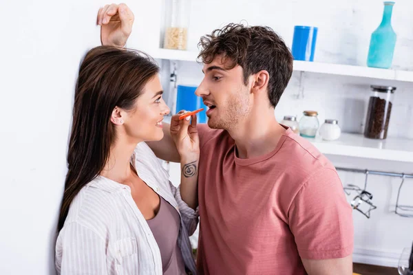 Smiling woman feeding boyfriend with slice of paprika near wall in kitchen — Stock Photo