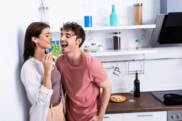 Mulher alegre segurando alface perto namorado com boca aberta na cozinha — Fotografia de Stock