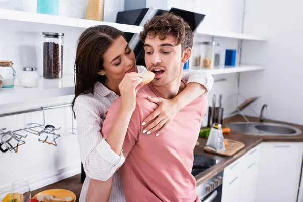 Jovem mulher abraçando e alimentando namorado com baguete na cozinha — Fotografia de Stock