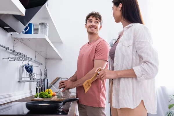 Uomo sorridente che tiene grattugia vicino a verdure e fidanzata con spatola vicino stufa in primo piano sfocata — Foto stock