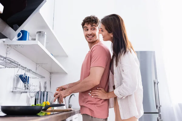 Seitenansicht einer lächelnden Frau, die ihren Freund umarmt, Reibe neben Gemüse hält und Pfanne auf Herd auf verschwommenem Vordergrund — Stockfoto