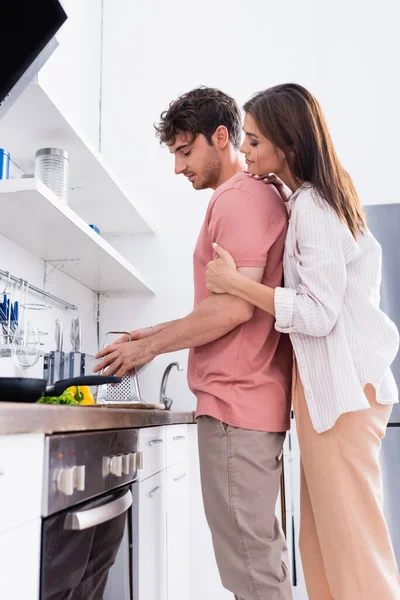 Side view of woman hugging boyfriend holding grater near vegetables in kitchen — Stock Photo