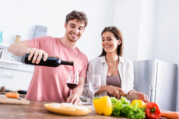 Uomo sorridente versando vino vicino fidanzata con coltello e verdure su primo piano sfocato — Foto stock