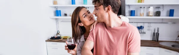 Brunette femme étreignant petit ami tout en tenant un verre de vin dans la cuisine, bannière — Photo de stock