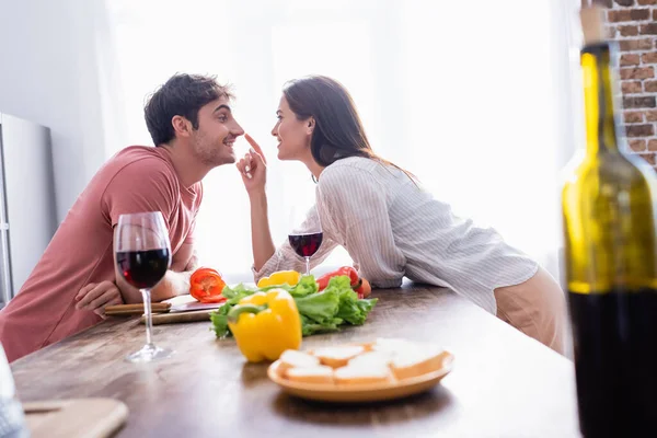 Smiling woman touching nose of boyfriend near wine and vegetables on blurred foreground — Stock Photo