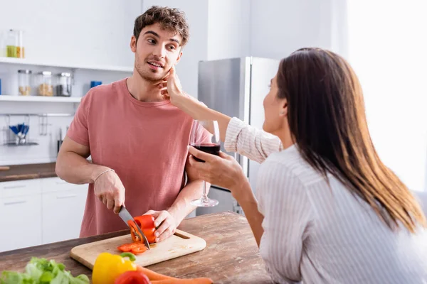 Young woman with glass of wine touching boyfriend cutting paprika near vegetables on blurred foreground — Stock Photo