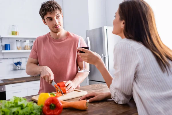 Young man cutting paprika and looking at girlfriend with glass of wine on blurred foreground — Stock Photo