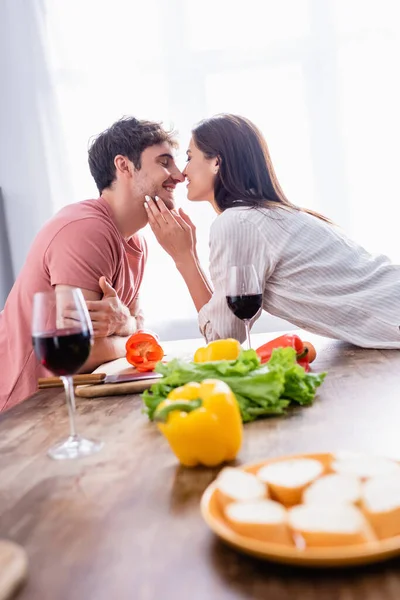 Smiling woman kissing boyfriend near vegetables and glasses of wine on blurred foreground — Stock Photo