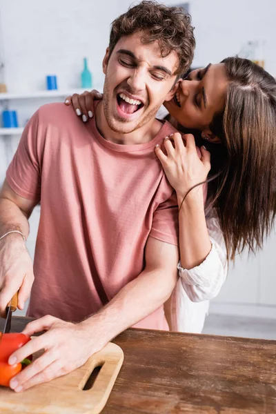 Усміхнена жінка обіймає хлопця, що ріже паприку на кухонний стіл — стокове фото