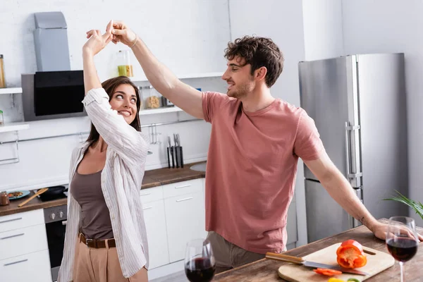 Hombre sonriente bailando con su novia cerca del pimentón y el vino en primer plano borroso en la cocina - foto de stock