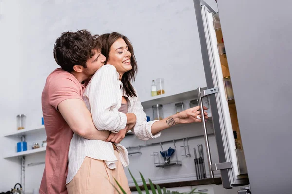 Hombre abrazando novia sonriente cerca de nevera abierta en la cocina - foto de stock