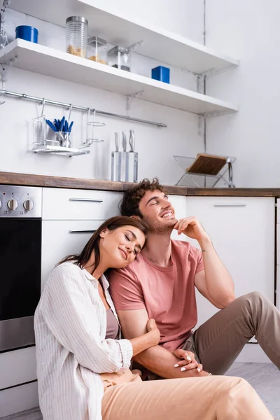 Smiling woman with closed eyes sitting near boyfriend on floor in kitchen — Stock Photo