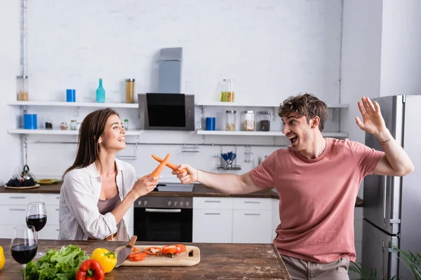 Cheerful couple fighting with carrots near vegetables and wine — Stock Photo