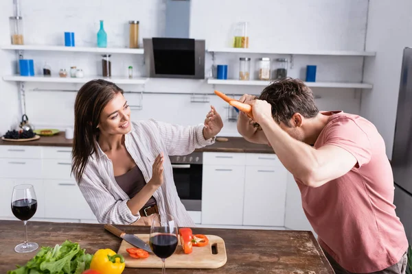 Mann hält Möhren neben lächelnder Frau, Gemüse und Wein auf Küchentisch — Stockfoto