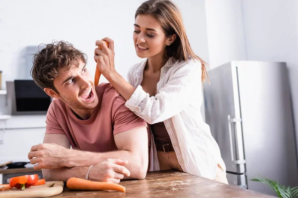 Lächelnde Frau hält Karotte neben aufgeregtem Freund in Küche — Stockfoto