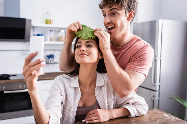 Sorrindo homem segurando alface perto da namorada tomando selfie em primeiro plano borrado — Fotografia de Stock