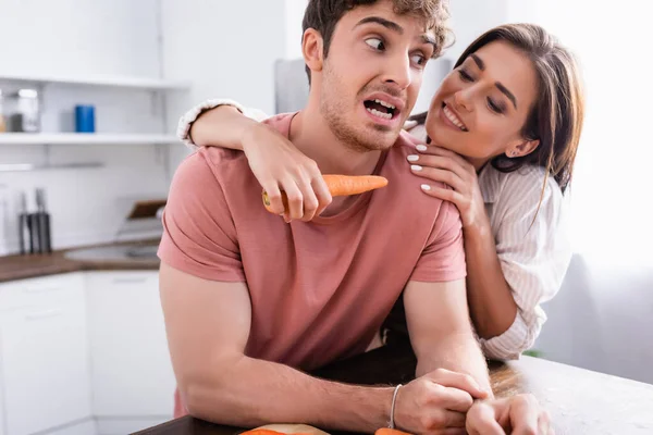 Femme souriante tenant la carotte près du petit ami effrayé dans la cuisine — Photo de stock