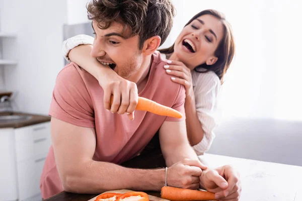 Мужчина кусает руку веселой девушки, держащей морковку на кухне. — стоковое фото