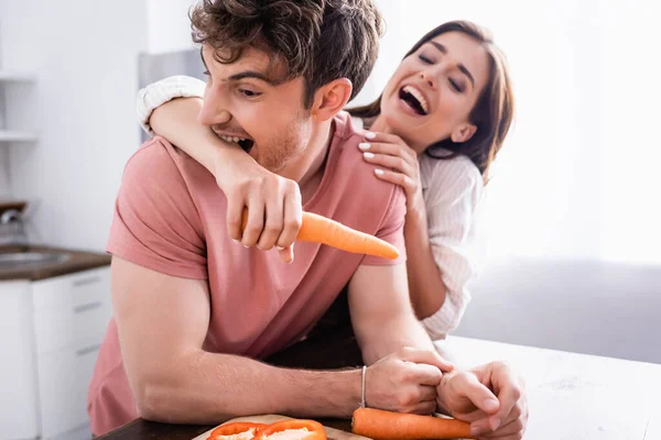 Man biting hand of cheerful girlfriend holding carrot in kitchen — Stock Photo