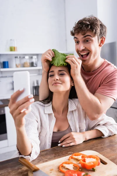 Homme joyeux tenant laitue près de petite amie avec smartphone prendre selfie dans la cuisine — Photo de stock