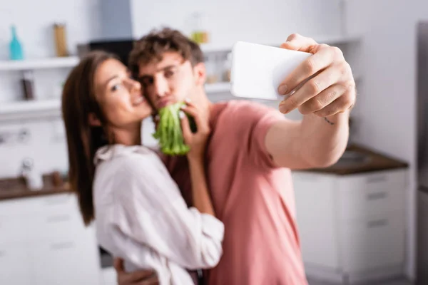 Смартфон в руке молодого человека, делающего селфи возле подружки с салатом на размытом фоне — стоковое фото