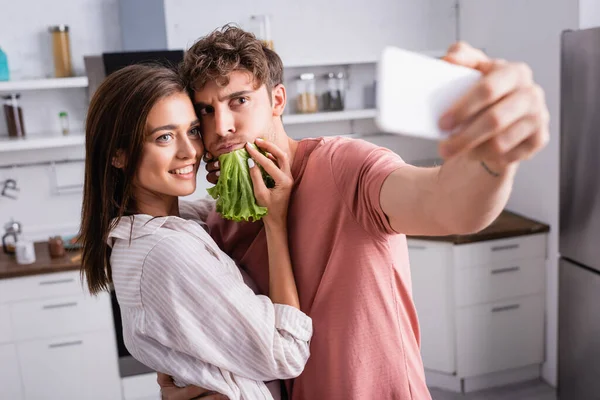 Smiling woman holding lettuce near boyfriend taking selfie on smartphone on blurred foreground — Stock Photo