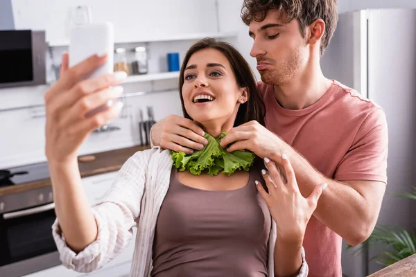 Man holding lettuce near smiling girlfriend taking selfie with smartphone on blurred foreground — Stock Photo
