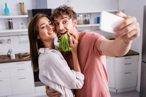 Cheerful couple with lettuce taking selfie on smartphone on blurred foreground — Stock Photo