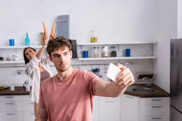 Homem tomando selfie com smartphone perto de namorada alegre com cenouras no fundo borrado — Fotografia de Stock