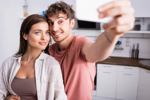 Smiling couple taking selfie with smartphone on blurred foreground in kitchen — Stock Photo