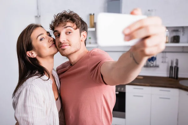 Sorrindo mulher de pé perto namorado tomando selfie no smartphone em primeiro plano desfocado — Fotografia de Stock