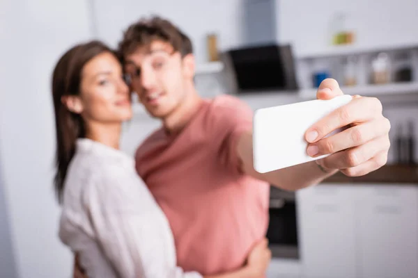 Smartphone in hand of man taking selfie near girlfriend on blurred background at home — Stock Photo