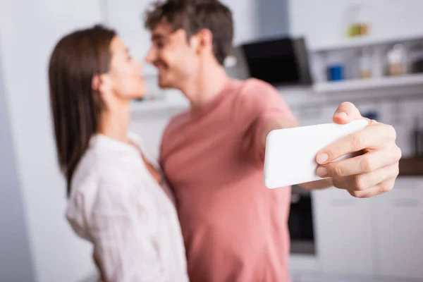 Smartphone in hand of man taking selfie while looking at girlfriend on blurred background — Stock Photo