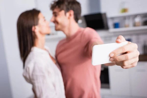 Smartphone in der Hand von Mann macht Selfie, während er Freundin auf verschwommenem Hintergrund ansieht — Stockfoto