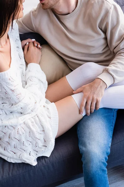 Vista recortada de la mujer joven en suéter de punto y calcetines de rodilla sentado cerca de novio en el sofá - foto de stock