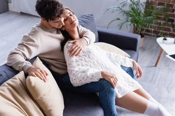 Young man hugging smiling woman in sweater on sofa with pillows — Stock Photo